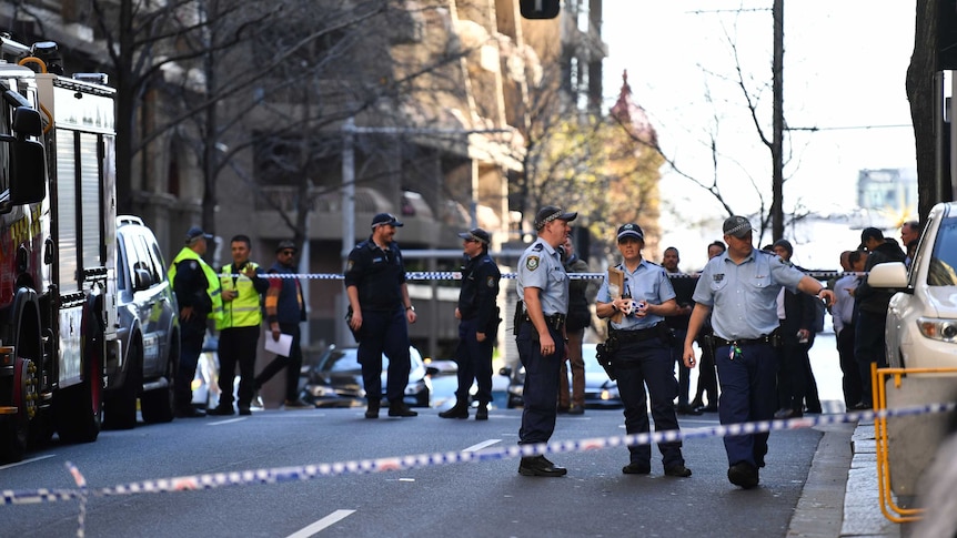 Police stand between emergency vehicles in a Sydney street, behind police tape.