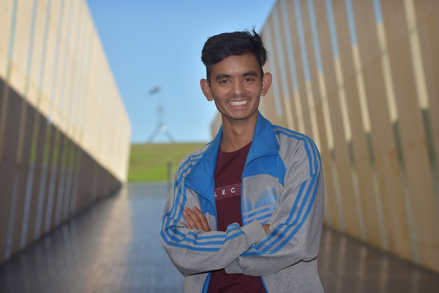 A man wearing a blue and grey jumper is smiling and crossing his arms with parliament house in the background