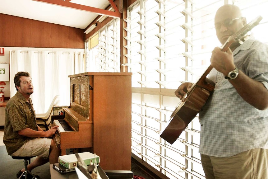 Man with finger on key of upright wooden piano watching man standing playing guitar before louvres