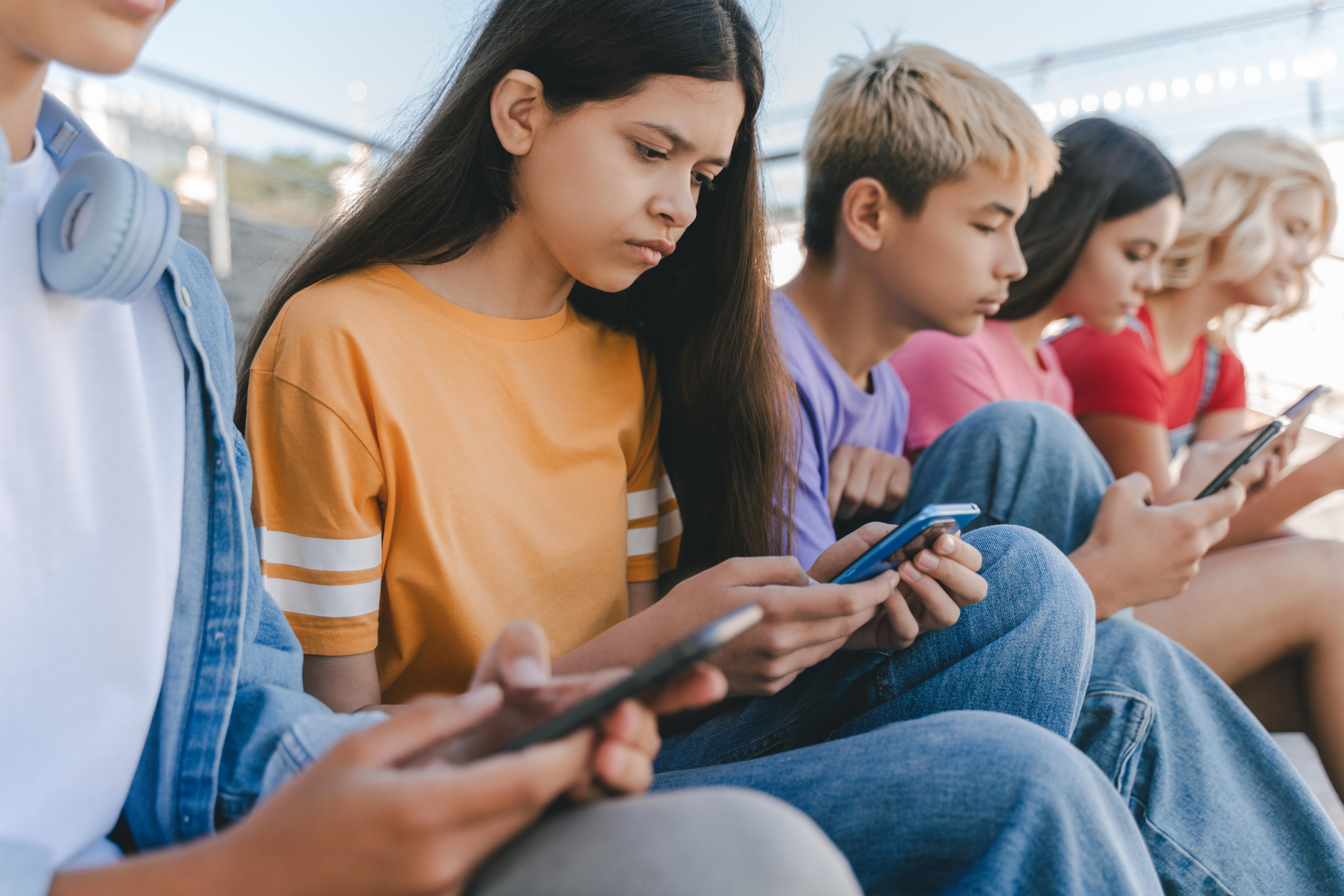 Five serious teenagers sitting in a row outside and using mobile phones.
