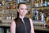 A woman wearing a black top sits in front of a restaurant bar, smiling at the camera.