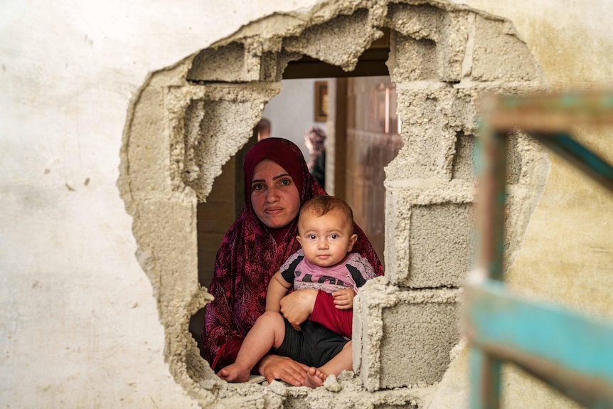 A woman holding a baby looks out a hole in the wall 