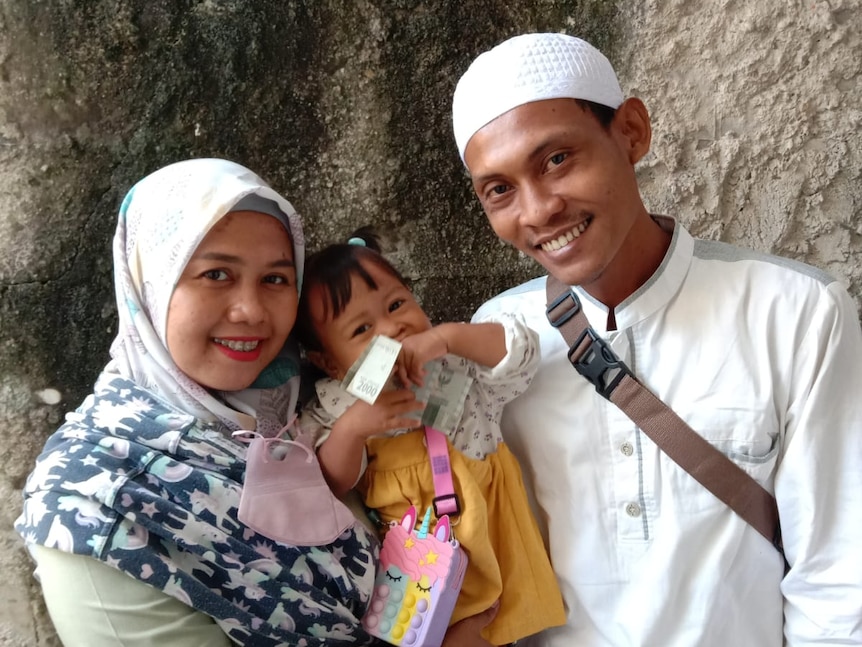 A family picture of three, parents wear muslim clothes, with a toddler in the middle, looking at the camera smiling,