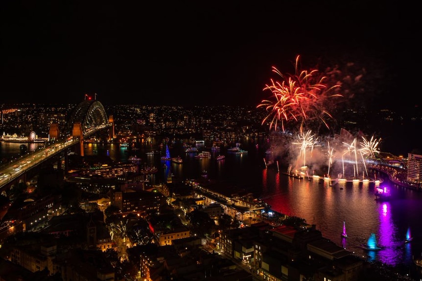An aerial view of Sydney Harbour and the Sydney Harbour Bridge at night time as orange and white fireworks explode in the sky.