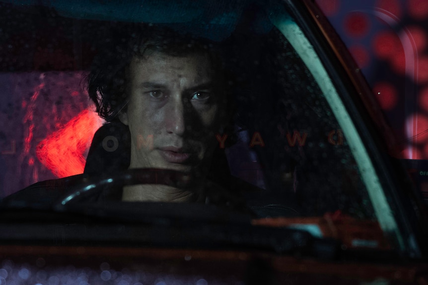 A middle-aged man in 80s-style dress looks bemused, behind a wheel at night. A motel sign can be seen outside the car. 