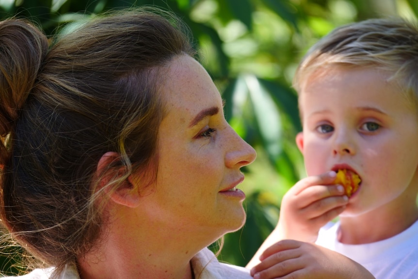 Close up on mum, profile and toddler eating fruit beside her shoulder in front of green in the garden.