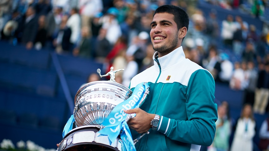 A tennis player wearing a green and white jacket holds a big silver trophy