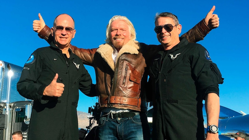 a man in a jacket raises his thumbs up next to two men in pilot uniforms