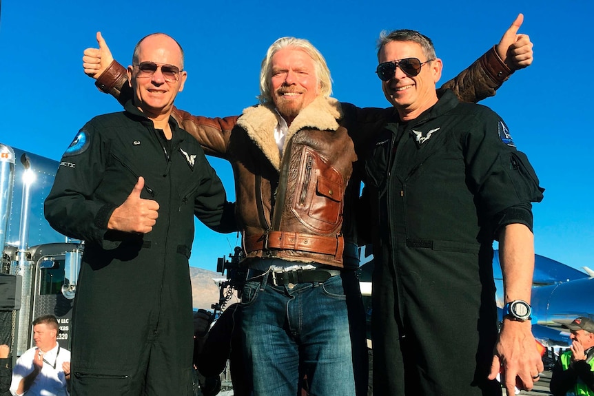 Richard Branson, wearing a jacket, raises his thumbs up next to two men in pilot uniforms