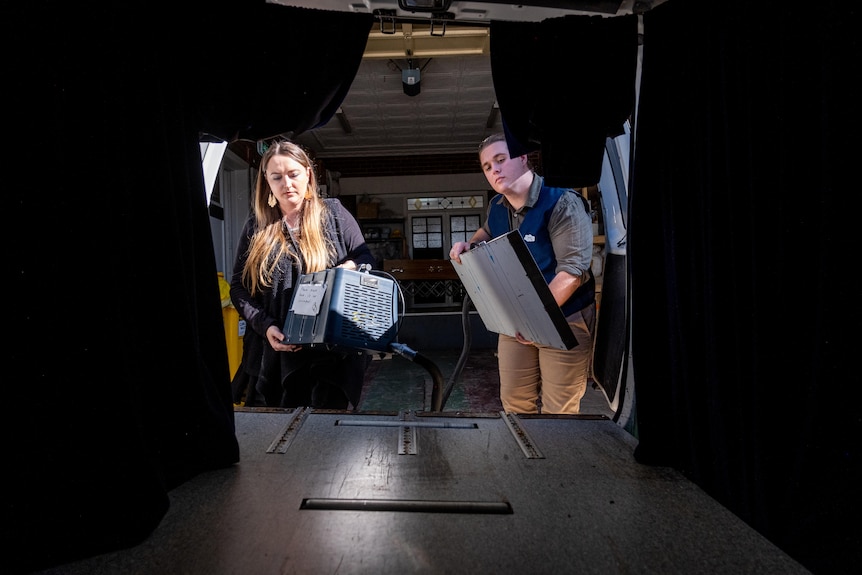 A man and a woman carry a stainless steel cold plate in a funeral service garage where the roller door is open