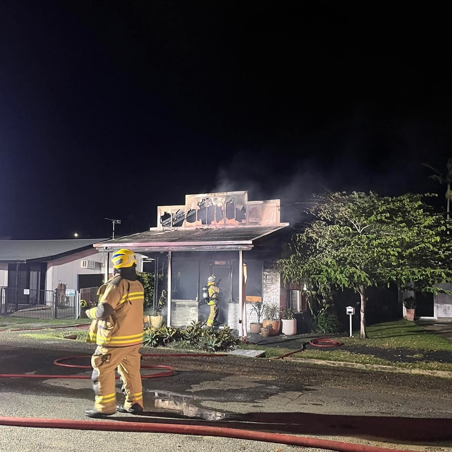 Fire crews in front of burnt building at night, cottage property, trees and grass 