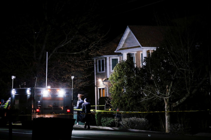 The exterior of a house lit up by the lights of an ambulance with people walking by