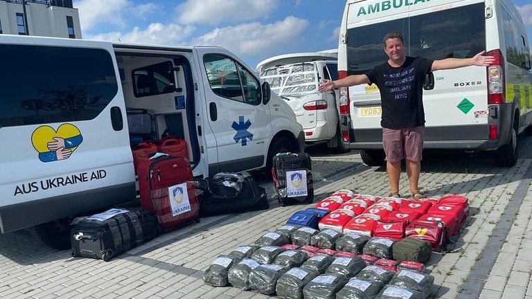 James Spenceley stands with his arms outstretched behind medical supplies and vehicles.