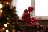 Two small children stare out the window wearing Christmas outfits