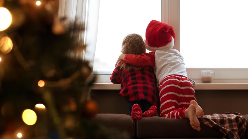 Two small children stare out the window wearing Christmas outfits