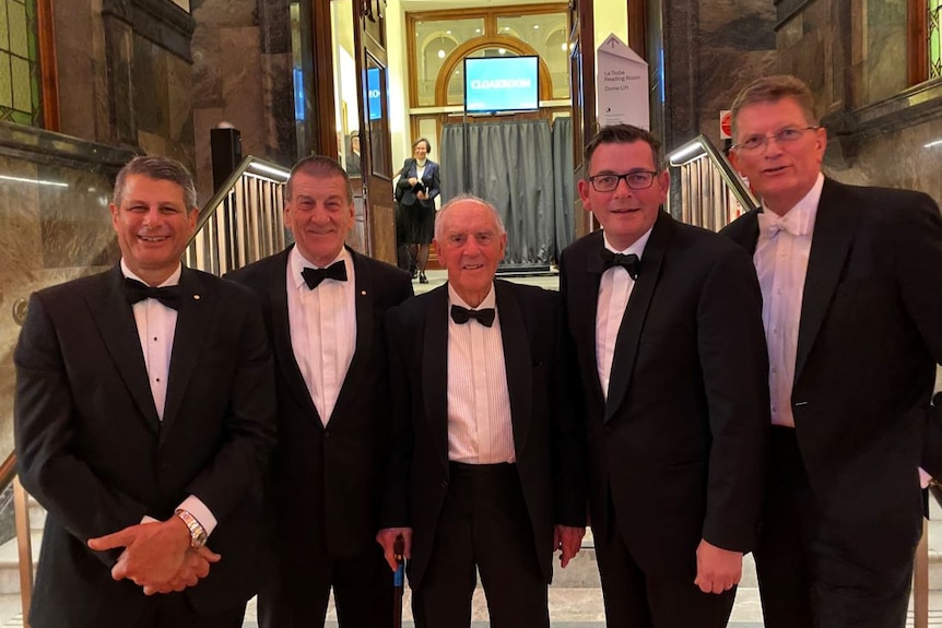 Five men in tuxedoes stand on marble steps.