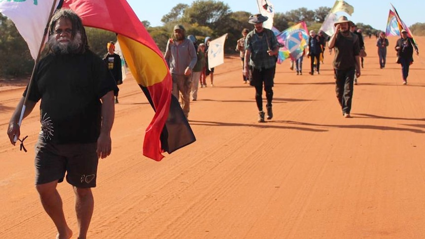 Kado Muir walks along a red dirt track holding an Aboriginal flagpole over his shoulder with other protestors walking behind him