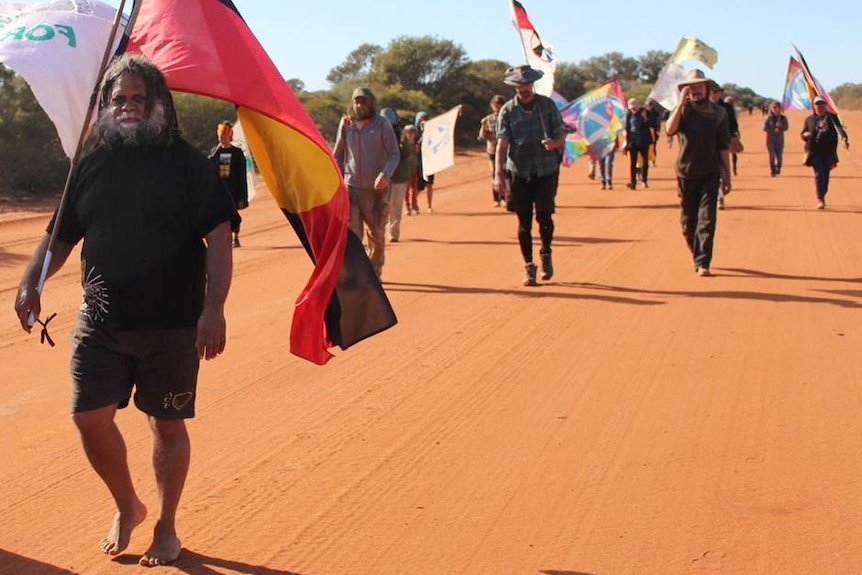 Kado Muir walks along a red dirt track holding an Aboriginal flagpole over his shoulder with other protestors walking behind him