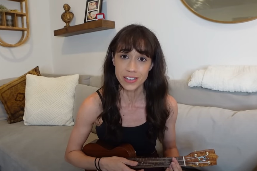 A screenshot of a woman sitting down and playing a ukulele