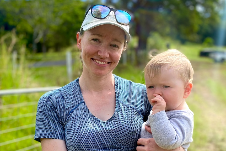 a woman stands smiling looking at the camera with her infant son