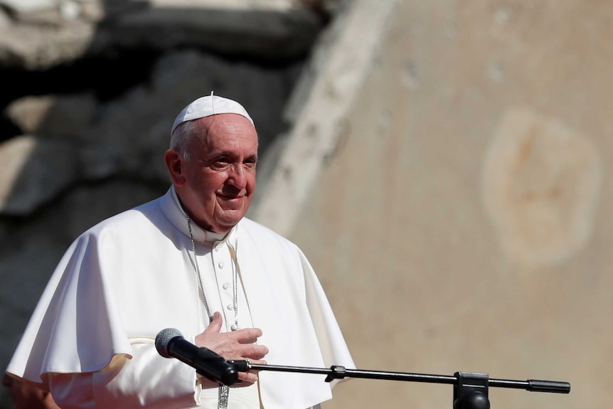The Pope smiles and touches his stomach with his right hand with damaged buildings behind him.