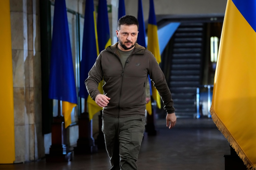Ukrainian President Volodymyr Zelenskyy walks through a hallway lined with large Ukrainian flags.