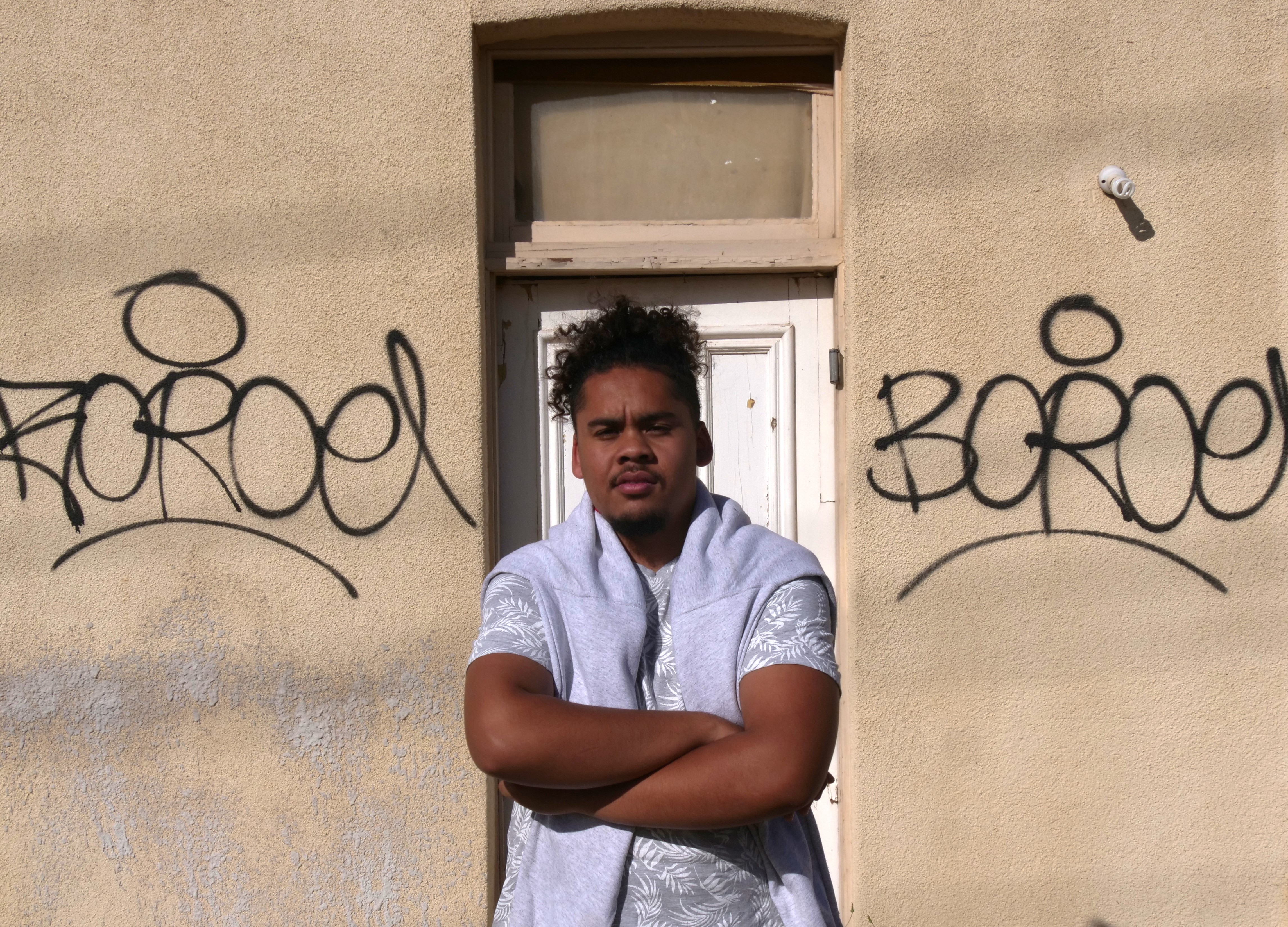 A young man in a white shirt stands in front of a wall with graffiti tags on it. 