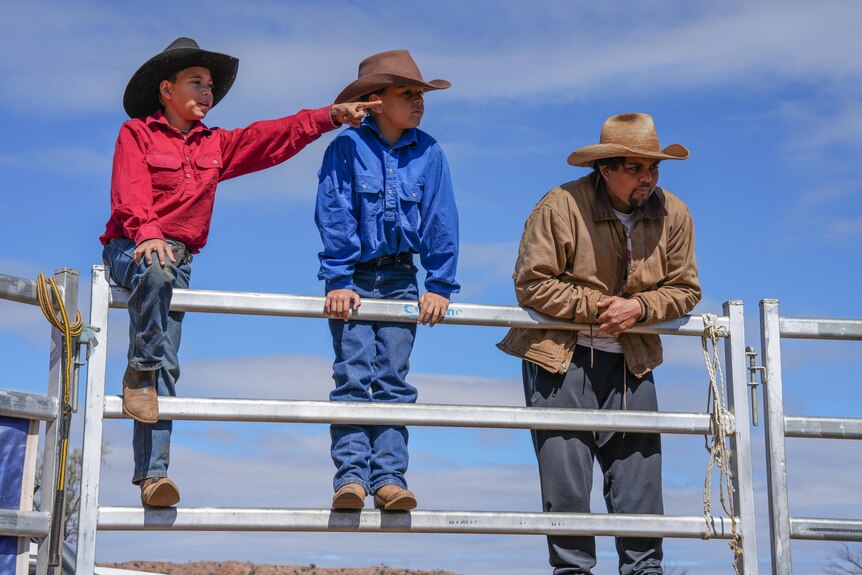Two young boys and a man dressed in stockmen gear lean on a metal cattle fence.