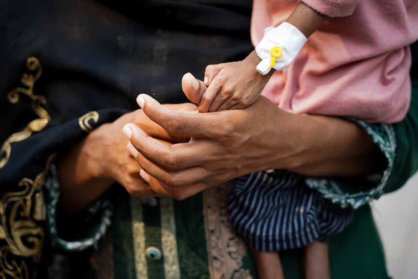 A child's tiny malnourished hand, with a catheter on his wrist, grips his mother's thumb as she holds him up