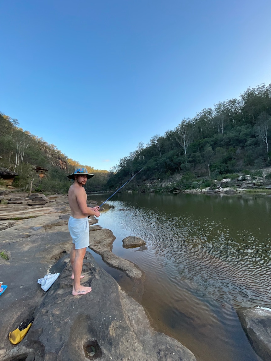 Harley Windsor standing on a rock beside a rocky creek and fishing with a rod. He wears a wide-brimmed hat and boardshorts.