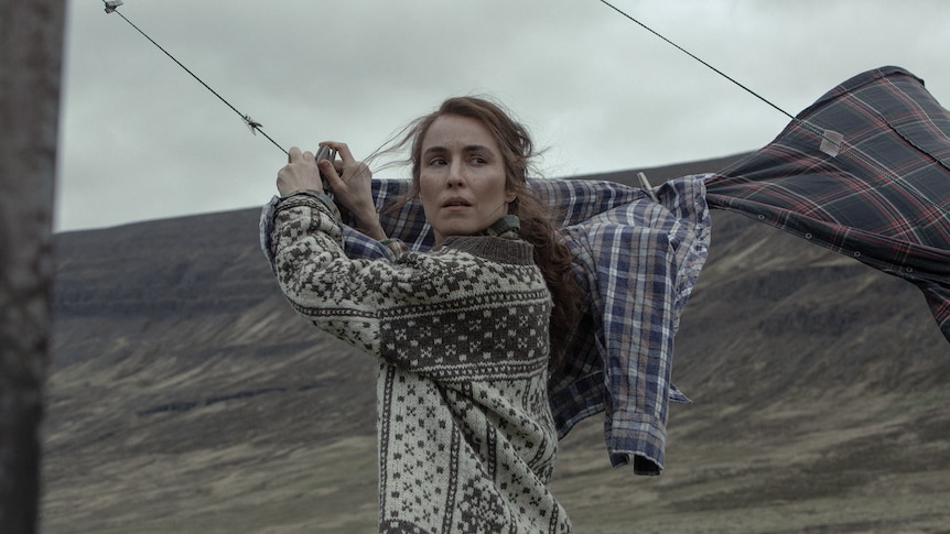 Woman with whispy brown hair, wearing a brown and white patterned cardigan hangs clothes on a line against overcast landscape
