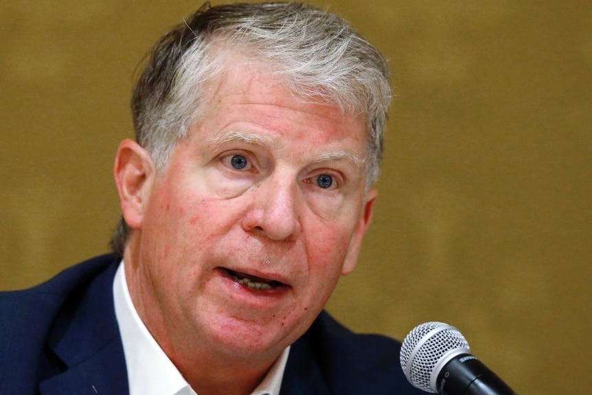 Cyrus Vance speaks into a microphone in front of a brown background.