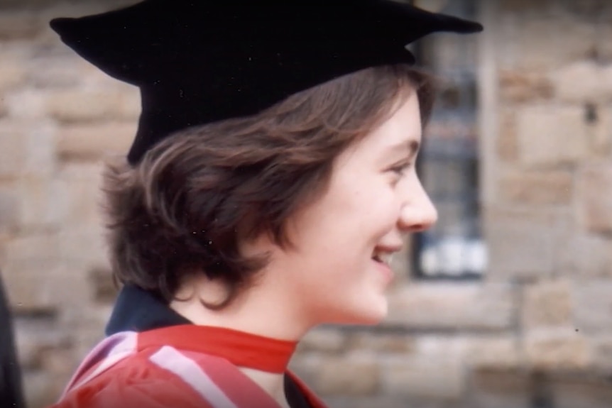 A young woman with short brown hair wears a graduate gap and gown, smiling.