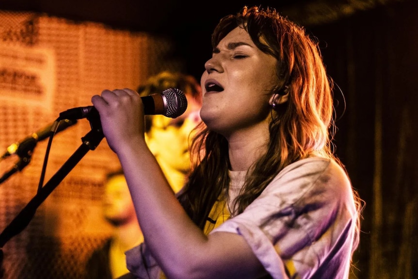 A young woman singing passionately into a microphone. She is wearing a tie-died t-shirt slightly rolled up at the sleeves. 