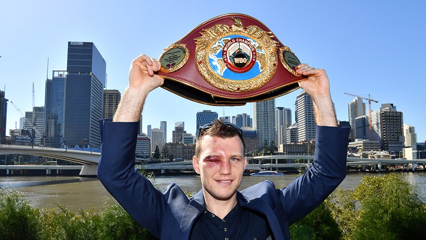 A man smiling as he holds a boxing title belt over his head.