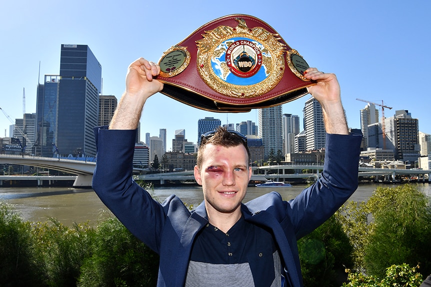 A man smiling as he holds a boxing title belt over his head.