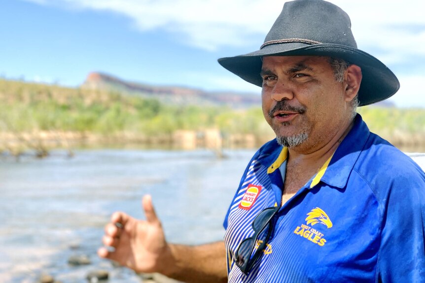 A man wearing a blue t-shirt and wide-brimmed hat stands next to a river.