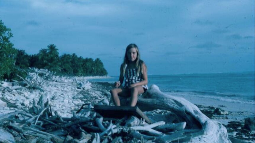 An old photograph of a girl with long, blonde hair sitting on a beach littered with driftwood.