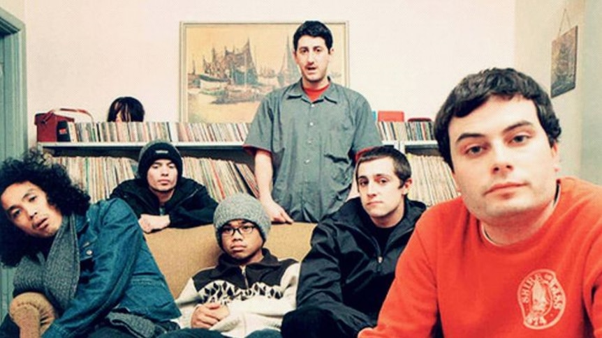 The six members of The Avalanches sitting on a couch in front of a shelf full of records and CDs