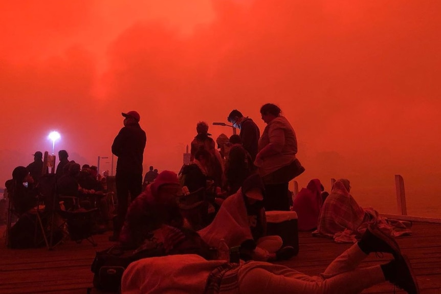 People seek refuge at Mallacoota Wharf under an orange sky.