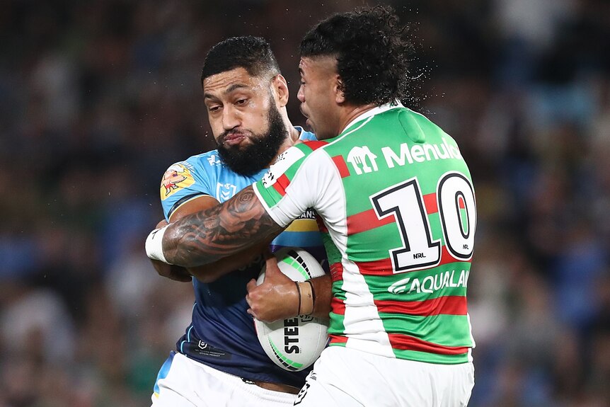 A Gold Coast Titans NRL player holds the ball as he is tackled by a South Sydney Rabbitohs opponent.