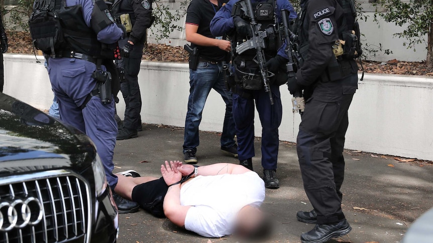 A handcuffed man laying face down on the ground surrounded by armed police
