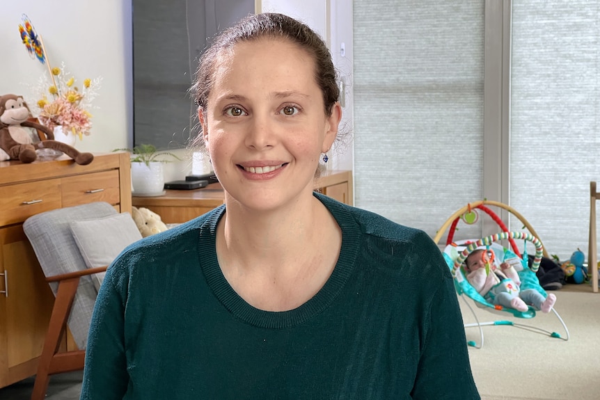 A woman smiles at the camera from her living room, her baby in a rocker and a monkey toy visible in the background.