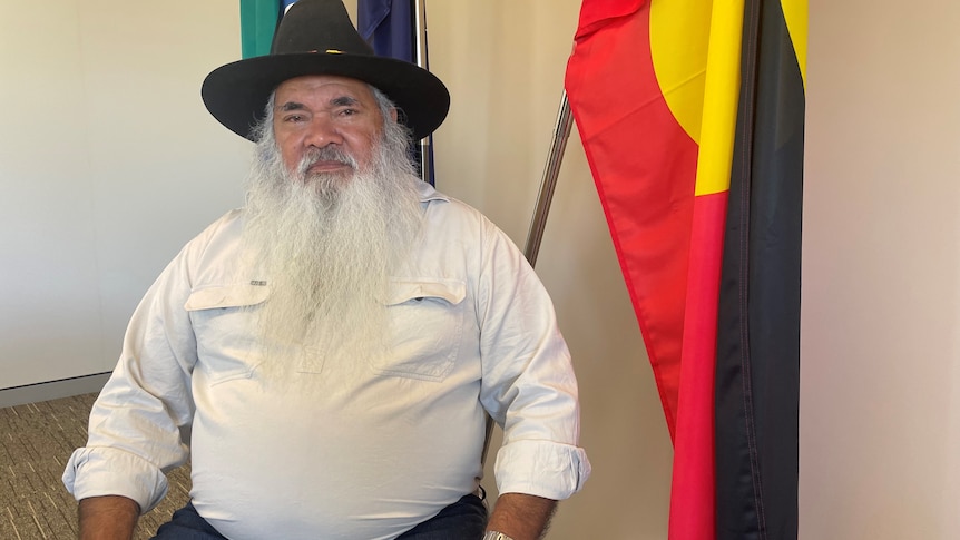Labor senator Pat Dodson wearing a black hat and sitting in front of an Aboriginal and a Torres Strait Islander flag.