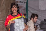 An Aboriginal woman holds her birth certificate in front of a researcher on a computer and sign saying ntscorp