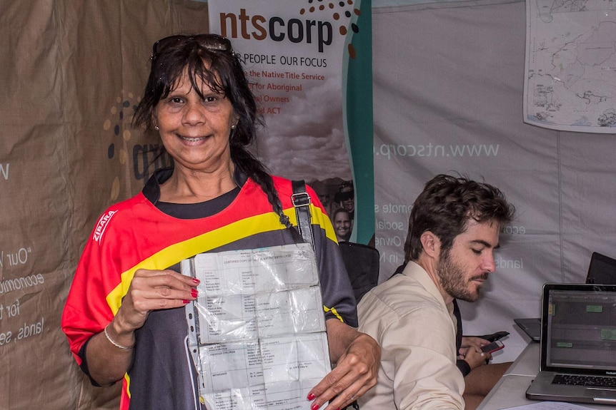 An Aboriginal woman holds her birth certificate in front of a researcher on a computer and sign saying ntscorp