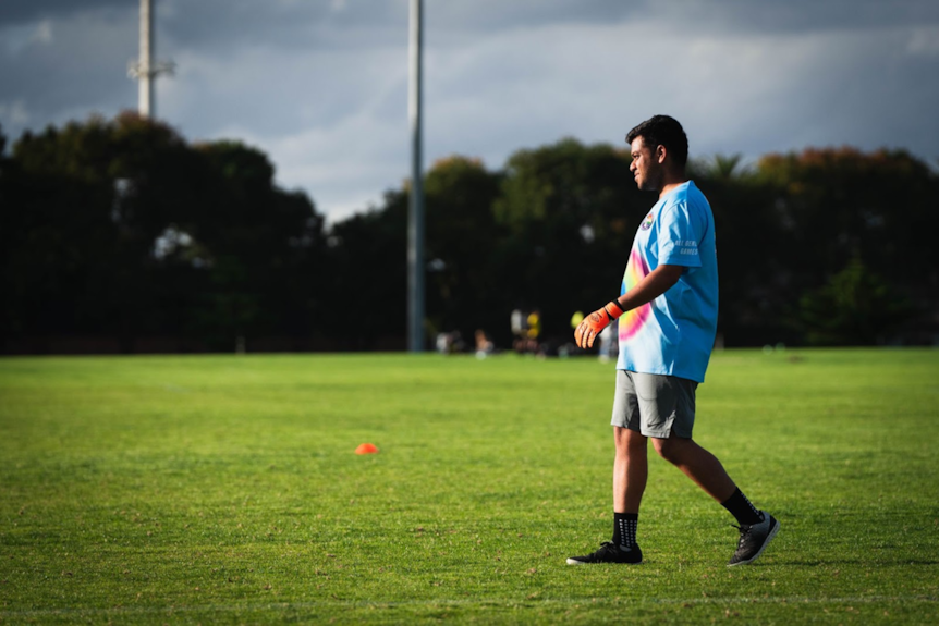 A man wearing a blue shirt stands on a football field, he is looking into the distance, pictured right of frame. 