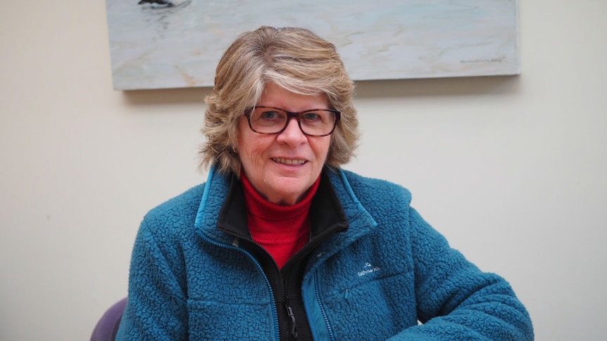 A woman with short blond hair and glasses sits at a table in a cafe.