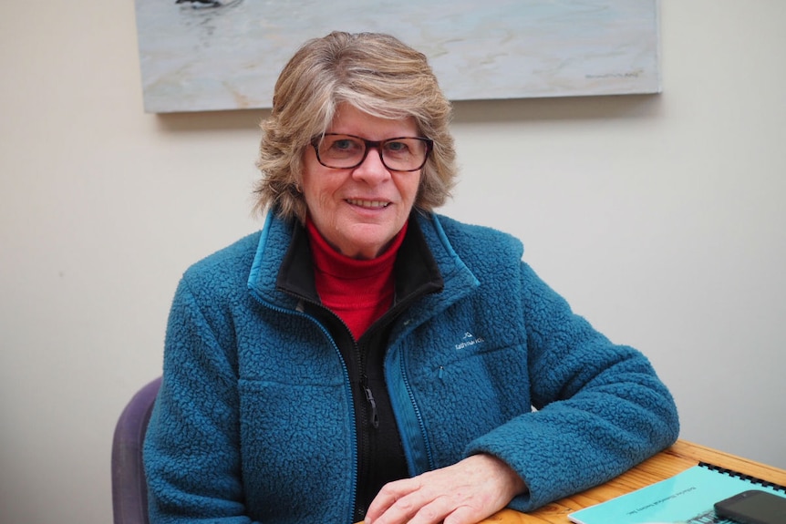 A woman with short blond hair and glasses sits at a table in a cafe.