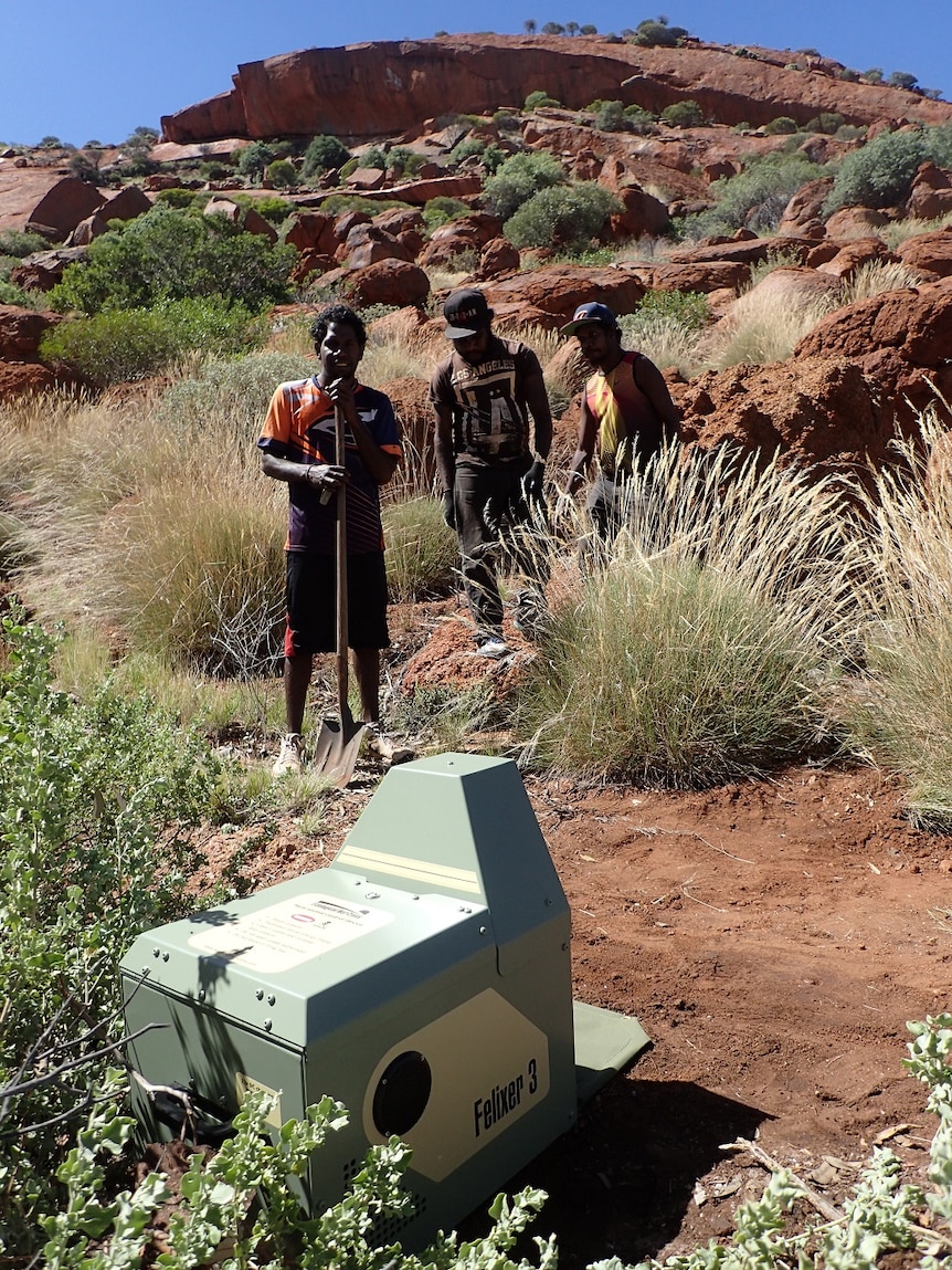 Three Aboriginal men standing in front of a rocky outcrop with a green knee-height machine in front of them.